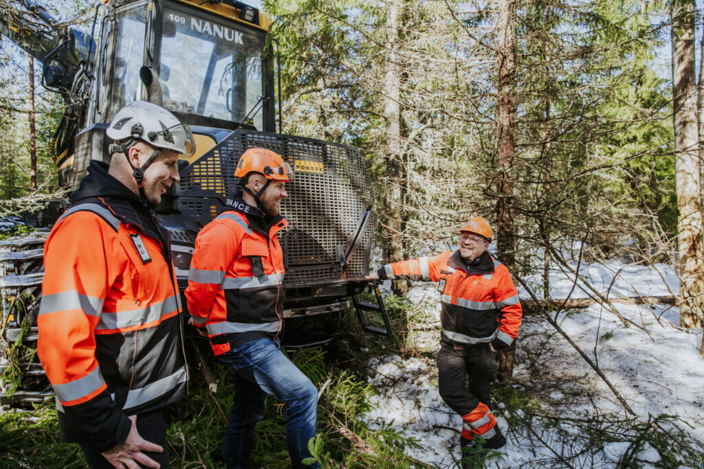 Metsäkoneen edessä kolme hymyilevää miestä työvaatetuksessa.